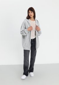 Sweat à capuche gris zippé avec une coupe décontractée, associé à un léger pull, un jean noir et des baskets blanches. Design simple, texture douce.