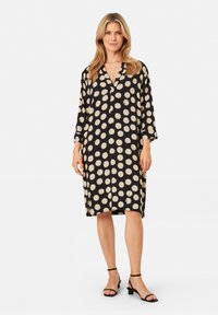 Schwarzes Kleid mit großen beigen Pünktchen, 3/4-Ärmeln, einem V-Ausschnitt und einer lockeren Passform, ergänzt durch schwarze Sandalen mit Knöchelriemen.