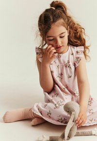 Rosa Blumenkleid mit Rüschensäumen und Spitzenbesatz am Saum. Kind trägt hellrosa Socken und hält ein beiges Stoffspielzeug. Neutraler Hintergrund.