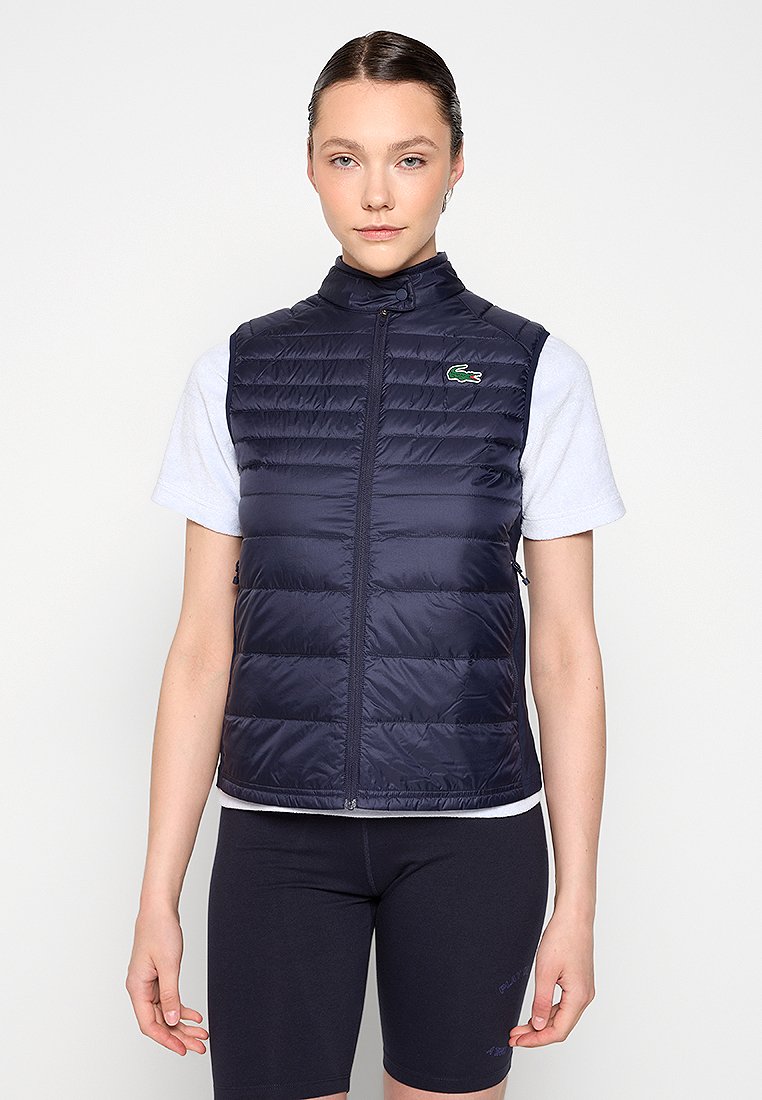 Jeune femme avec des cheveux plaqués en arrière portant un gilet matelassé navy par-dessus une chemise blanche à manches courtes et un short de cyclisme noir, se tenant contre un fond blanc.