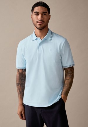 Mann mit kurzen dunklen Haaren und Tätowierungen auf beiden Armen, trägt ein hellblaues Poloshirt und dunkle Hose, steht mit einer Hand in der Tasche.