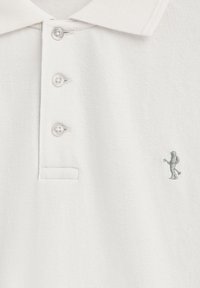 Camisa polo blanca con tejido texturizado, tres botones y una pequeña figura bordada en el lado izquierdo.