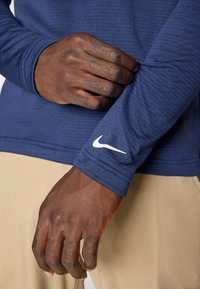 Granatowa sportowa koszulka z długim rękawem o teksturowanym materiale, z białym logo Nike na mankiecie. Dłoń trzymająca rękaw pokazuje tatuaż.