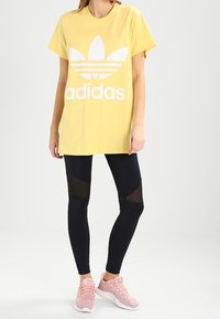 Yellow cotton t-shirt with white Adidas logo; paired with black leggings featuring mesh paneling; pink athletic shoes with textured design.