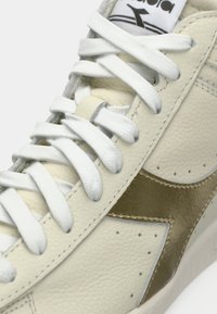 Baskets en cuir blanc avec un accent métallique doré sur le côté, surface texturée et lacets blancs. Design classique avec des détails perforés.