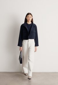 Femeie mergând în interior, purtând o jachetă neagră scurtă, pulover bleumarin, pantaloni albi largi, pantofi bej și având o geantă mică neagră de mână.