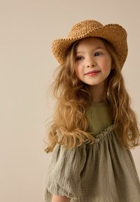 Un chapeau de cowboy en paille beige sur un enfant aux longs cheveux ondulés, portant une robe vert olive clair avec un buste froncé et des manches bouffantes.
