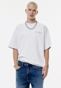 Ung person med kort blond hår iført en lys grå oversized T-shirt, blå jeans, sølv kæderekæde og armbånd mod en hvid baggrund.