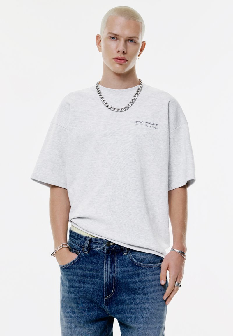 Junge Person mit kurzem blonden Haar, die ein hellgraues, übergroßes T-Shirt, blaue Jeans, eine silberne Kettenkette und Armbänder vor einem weißen Hintergrund trägt.