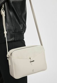 Kremowa torebka crossbody z teksturowanej ekoskóry, zapinana na zamek, z ozdobnym paskiem. Kontrastowy panel z zamszu z przodu z metalicznym akcentem.