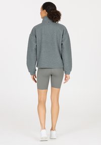 Grauer Fleece-Pullover mit hohem Kragen und strukturierter Oberfläche. Kombiniert mit hellgrauen Radshorts und weißen Turnschuhen.