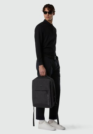 Uomo con camicia nera a maniche lunghe e pantaloni, occhiali da sole, sneaker bianche, che tiene uno zaino nero rettangolare North Sails per la maniglia superiore.