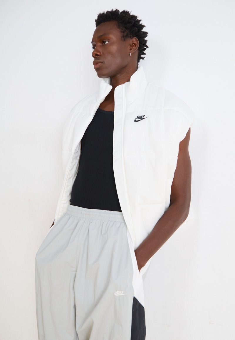Veste sans manches blanche en duvet avec un col montant et un logo Nike noir, associée à un pantalon gris et un débardeur noir.