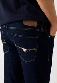 Jeans in denim blu scuro con cuciture gialle, dotati di una toppa in pelle con marchio e tasche posteriori angolate con un'etichetta a forma di triangolo.