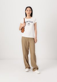 Maglietta grafica bianca con scritte blu, pantaloni beige a gamba larga e sneakers bianche. Piccola borsa marrone con tracolla tenuta in una mano.