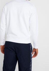 Vit sweatshirt med ribbad mudd och nederkant, i kombination med mörkblå byxor med ett upprepat logomönster längs sidan.