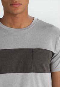 Camiseta de manga corta gris y gris oscuro con cuello redondo y un bolsillo en el pecho. Tejido de algodón con una textura suave.