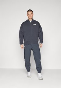 Dunkelgraue Bomberjacke mit Reißverschluss und weißem "FCBM"-Logo, kombiniert mit passenden Cargo-Hosen und weißen Sportschuhen. Glatte Textur, lässige Passform.