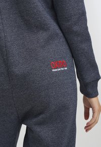 Grå hettegenser med teksturert finish. Har røde broderte detaljer på baksiden. Mansjettene er tettsittende, og stoffet ser mykt og komfortabelt ut.