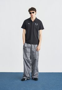 Νεαρός άνδρας που φοράει μαύρο πόλο BMW Motorsport, φαρδιά γκρι παντελόνια, μαύρα παπούτσια και στρογγυλά μαύρα γυαλιά ηλίου, στέκεται πάνω σε μπλε πάτωμα.