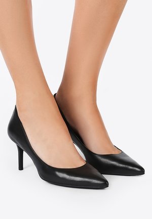 Pieds chaussés de talons hauts en cuir noir classique à bout pointu avec des talons aiguilles fins de hauteur moyenne sur fond blanc.