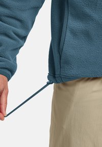 Fleece jack in het blauw, met een gestructureerde oppervlakte, elastische tailleband met een trekkoord, en een hand van een persoon die de pasvorm aanpast.
