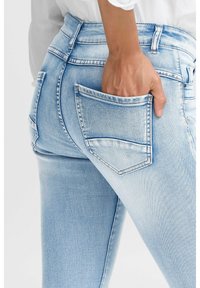 Jeans de denim azul claro con un diseño ajustado, cinco bolsillos y un ligero desvanecimiento. El bolsillo trasero está parcialmente cubierto por una mano.