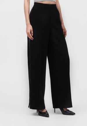 Donna che indossa pantaloni neri a vita alta e gamba larga con cerniere laterali, abbinati a scarpe nere con tacco alto e punta appuntita e top grigio corto.