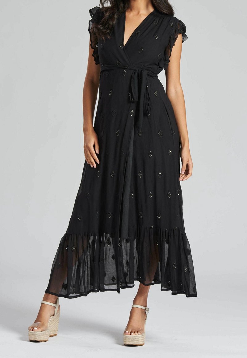 Schwarzes Wickelkleid mit RüSchlein, verziert mit silbernen Diamantelementen und einem gestuften Saum. Kombiniert mit leichten Keilabsätzen.