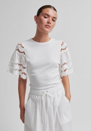 Woman wearing white short-sleeve top with lace cutouts on puffed sleeves and white drawstring pants, standing with one hand in pocket.