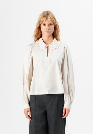 Blusa di crema con colletto arricciato e scollatura a chiave, caratterizzata da maniche lunghe a sbuffo e bordi scallop. Abbinata a pantaloni scuri.