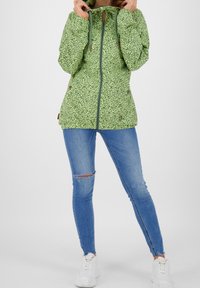 Zielona kurtka w wzory z kapturem, z przodu na zamek i sznurkami do regulacji. Ma luźny krój, dwa boczne kieszenie i gładką teksturę.