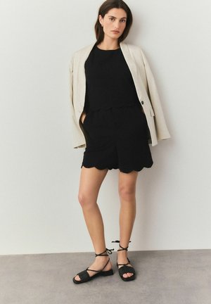 Haut noir à bords festonnés et shorts assortis, accompagnés d'un blazer beige. Sandales noires en conflit avec des liens à la cheville. Texture lisse, design minimaliste.