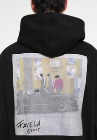 Fekete kapucnis pulóver, amelynek hátulján nagy grafika látható szürke és sárga színekben, emberekkel, akik egy autó mellett sétálnak, "Favela ©2015" aláírással.