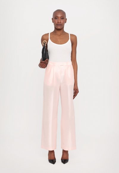 Pale pink wide-leg trousers with a smooth texture, pleated front, and a matching belt. Model holds a black handbag with a gold accent.