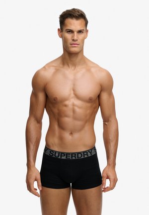 Muskelbepackter Mann, der frontal steht, trägt schwarze Superdry-Boxershorts, neutraler Gesichtsausdruck, einfarbiger heller Hintergrund.