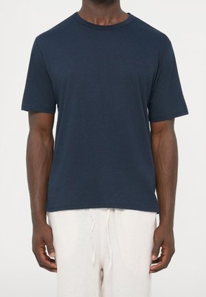 Homme portant un t-shirt bleu foncé uni à manches courtes et un pantalon beige clair à cordon, debout devant un fond blanc.