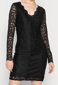 Robe en dentelle noire avec des manches longues et un décolleté en V. Présente des motifs floraux complexes et une silhouette ajustée, mettant en valeur un tissu texturé.