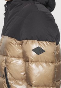 Gros plan sur une doudoune bicolore avec une section supérieure noire et une section inférieure bronze brillante, ornée d'un patch logo noir en forme de losange.