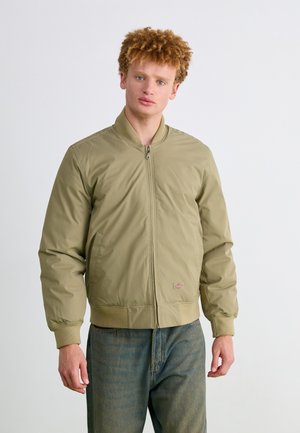 Chaqueta bomber verde oliva con cierre frontal, puños y dobladillo acanalados; llevada sobre pantalones de mezclilla. Presenta un pequeño logo en el pecho.