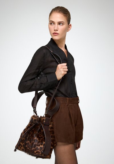 Torebka typu bucket w leopardziej, futrzanej estetyce z brązowymi skórzanymi akcentami, z zapięciem na sznurek i regulowanym paskiem. Modelka ma na sobie czarną bluzkę i brązowe szorty.