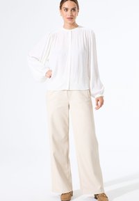 Witte blouse met lange mouwen, knoopsluiting en geplooide details, gecombineerd met beige wijde broek en bruine instappers. Eenvoudig en strak ontwerp.
