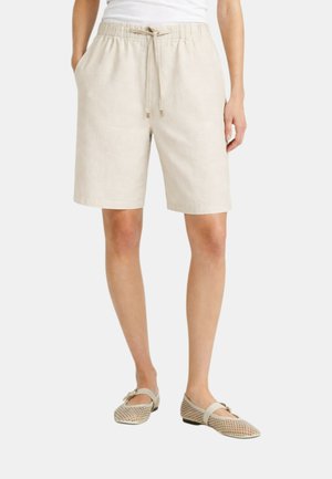 Persona che indossa pantaloncini beige con coulisse fino al ginocchio abbinati a ballerine Mary Jane in rete beige, in piedi davanti a uno sfondo semplice.