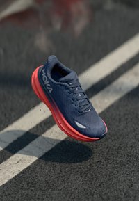 Baskets de course légères bleu vif avec un dessus en maille, semelle intermédiaire rouge, semelle texturée et accents réfléchissants. Logo HOKA affiché de manière proéminente.