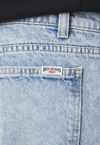 Svetlo modré džínsy majú zadnú kapsu s bielou nálepkou s nápisom "BDGJEANS," so žltým šitím a mierne textúrovaným povrchom.