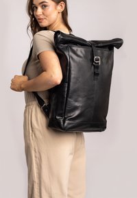 Sac à dos en cuir noir à rabat, avec des bretelles ajustables, un fermoir à boucle à l'avant et une texture lisse, porté sur l'épaule.