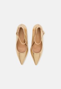 Pompes à talons hauts en métal doré avec des bouts pointus et une bride à la cheville. Texture lisse avec une finition légèrement brillante. Intérieur neutre.