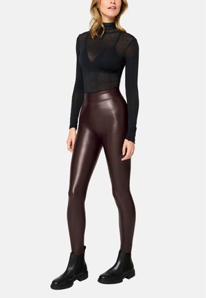Woman wearing sheer black long-sleeve top over black bralette, high-waisted shiny dark brown leggings, and black ankle boots, standing.