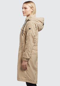 Beiger, langer Regenmantel mit Kapuze, elastischen Bündchen, Reißverschluss vorne und einer Brusttasche. Glattes, leichtes Material mit minimalen Nähten.