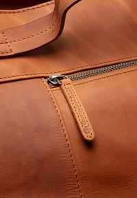 Bolso de cuero marrón con una cremallera y herrajes plateados, detalles de costura y una pequeña etiqueta de cuero adjunta cerca de la cremallera.
